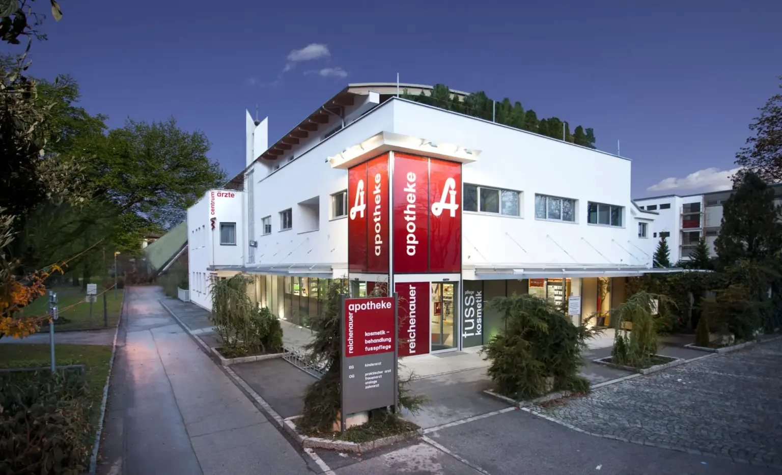 Weißes Gebäude der Reichenauer Apotheke in der Abenddämmerung mit rotem Turm „apotheke“ und Apotheken-A, davor das Hinweisschild